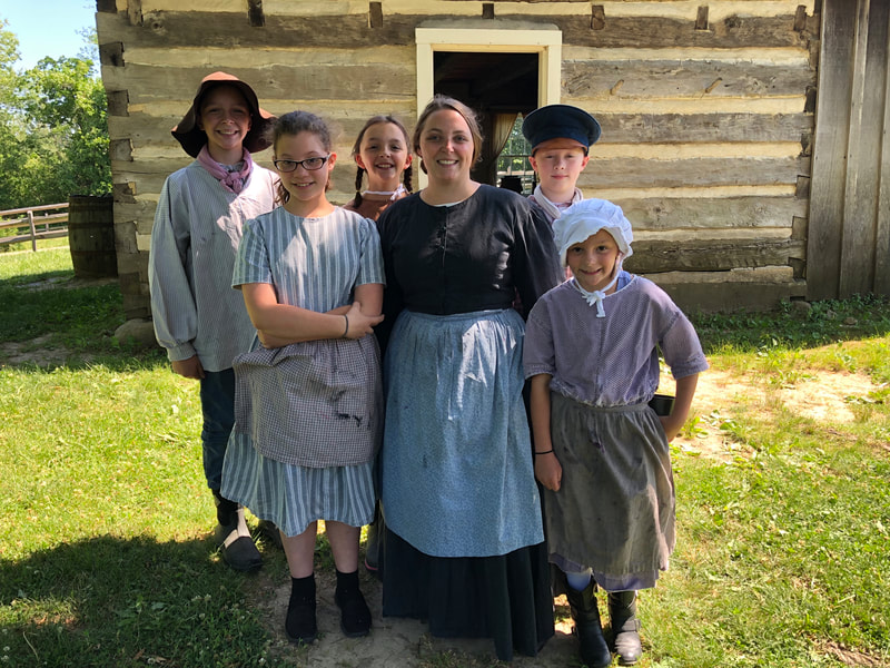 Ms. Boston is standing, smiling, with five campers. They are all dressed in 1840's historical clothing and there is a log cabin behind them.